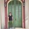 Two people stand near a green arched doorway on a light sidewalk, facing each other.