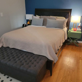 Bedroom with bed, beige bedding, blue wall, dark headboard, green nightstand, lamp, ottoman, wooden floor