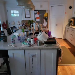 Cluttered kitchen island with dishes, cups, and cleaning supplies in a bright homey kitchen.