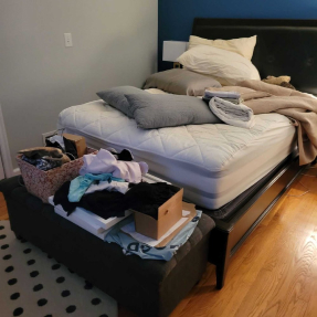 Messy bedroom with bare mattress, piled blankets, ottoman of laundry, and scattered items.
