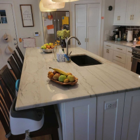 Modern kitchen with marble island, sink, fruit bowls, white cabinets, and stainless appliances.
