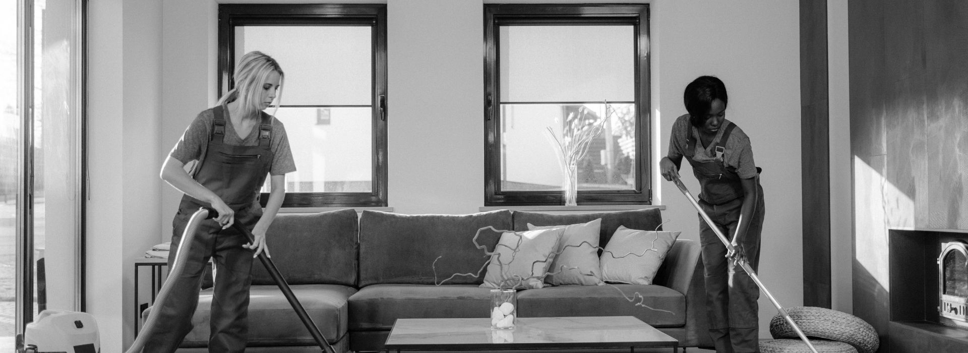 Two people in overalls vacuuming a living room with a sofa, coffee table, and fireplace (grayscale)