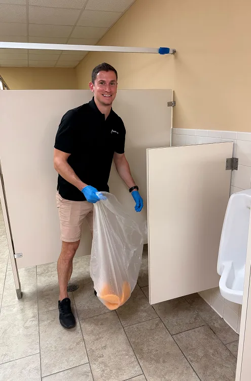 Man in black polo and shorts holding a large trash bag with orange contents in a restroom.