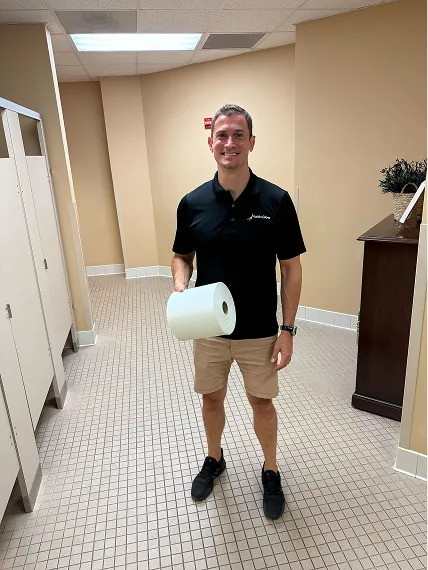 Man in black polo and tan shorts holding a roll of paper towels in a beige tiled room.