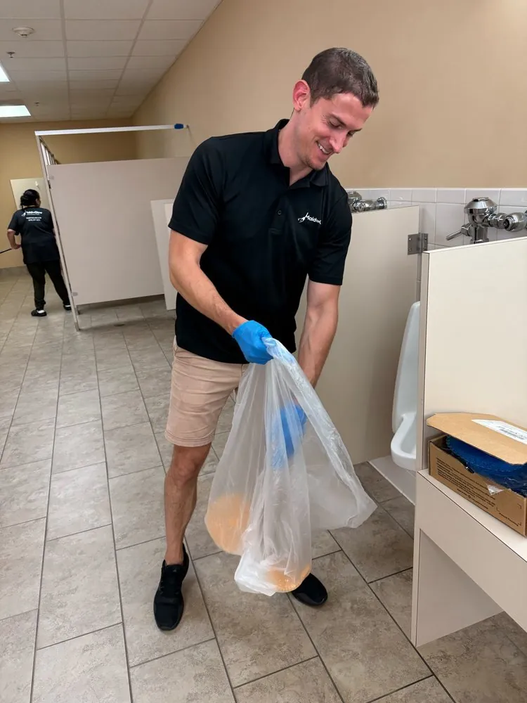 Man in black polo and beige shorts holding a plastic bag with two round orange-brown objects in a break room.