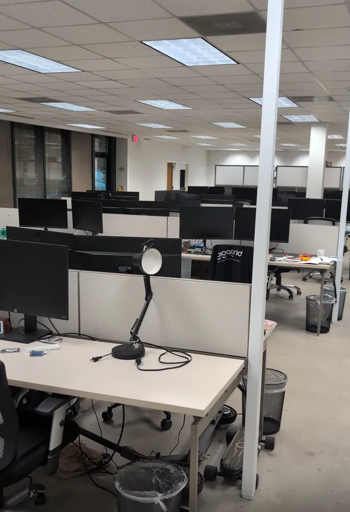Open-plan office with multiple workstations, monitors, desks, and a desk lamp in foreground.